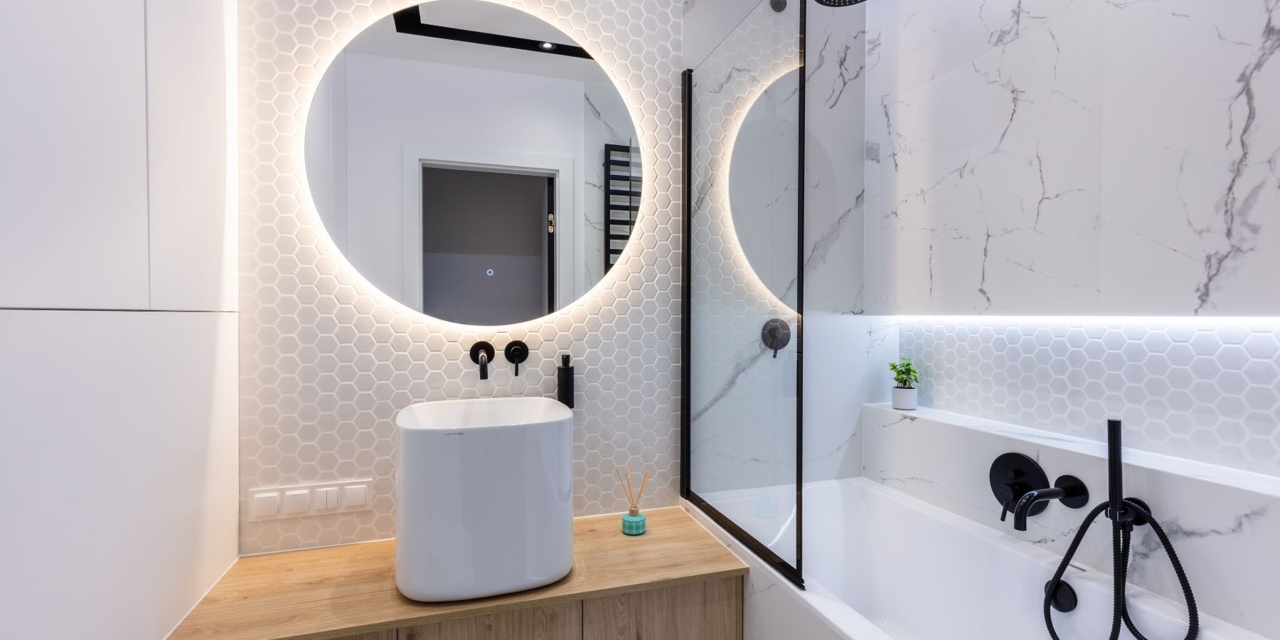 Modern bathroom with round illuminated mirror, vessel basin, bathtub, glass screen and black fittings